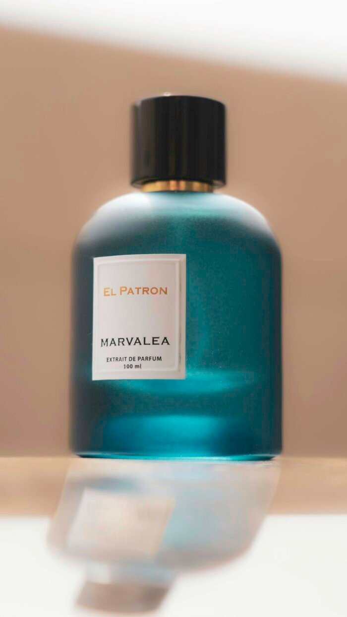 A chic teal perfume bottle labeled El Patron Marvalea, resting elegantly on a reflective surface.