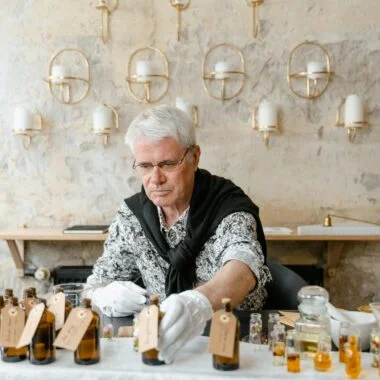 Elderly man crafting fragrances indoors, surrounded by essential oils in bottles.