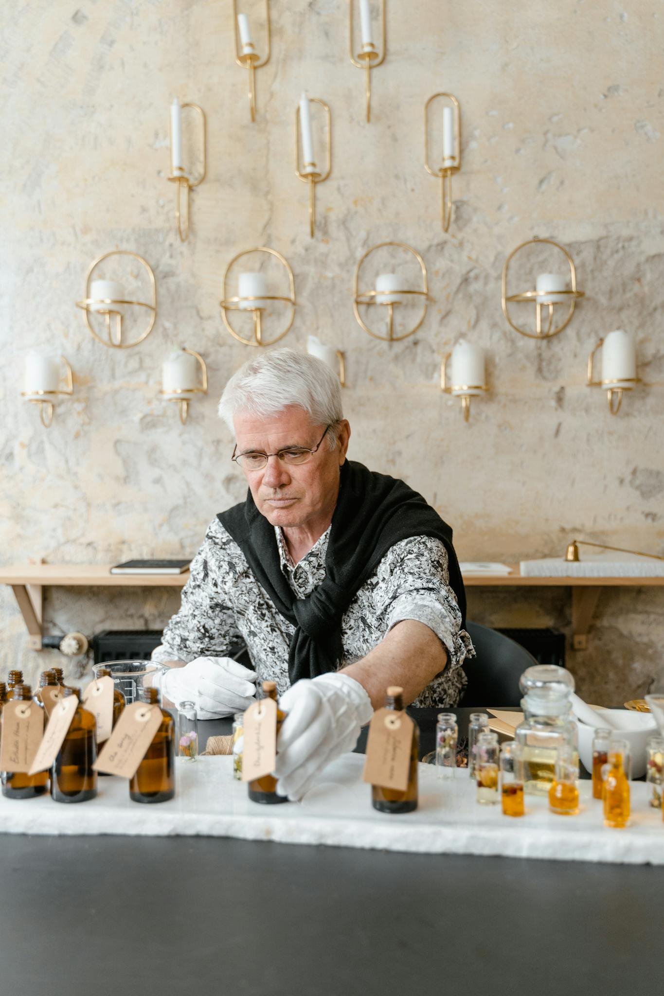 Elderly man crafting fragrances indoors, surrounded by essential oils in bottles.